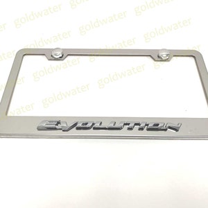 May include: Silver metal license plate frame with the word "EVOLUTION" in raised letters. The frame has two screw holes on the top and two on the bottom.