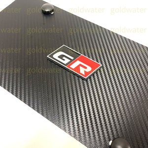 May include: Black carbon fibre license plate frame with a silver and red "GR" emblem.