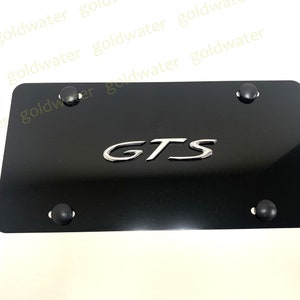 May include: A black license plate with the chrome letters "GTS" in the center. Four black rubber bumpers are placed at each corner. The plate is rectangular and has a glossy finish.