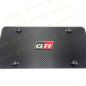 May include: Black carbon fibre license plate frame with a red and white "GR" logo. The frame has four black plastic screw covers.