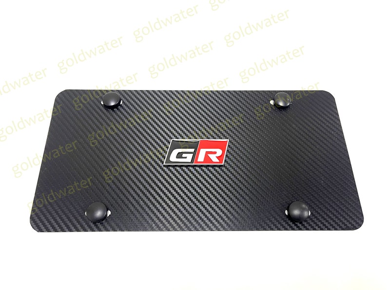 May include: Black carbon fibre license plate frame with a red and white "GR" logo. The frame has four black plastic screw covers.