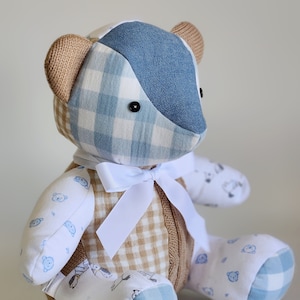 May include: A patchwork teddy bear made from blue and white gingham fabric, brown knitted fabric, and white fabric with a blue dog print. The bear has a white bow around its neck.