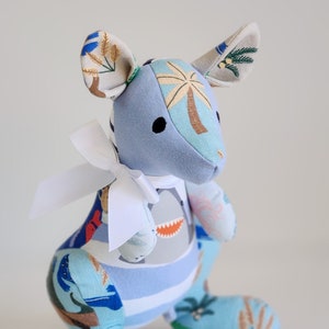May include: A blue and white patchwork stuffed kangaroo toy with a white bow. The kangaroo has a palm tree design on its head and a shark design on its belly.