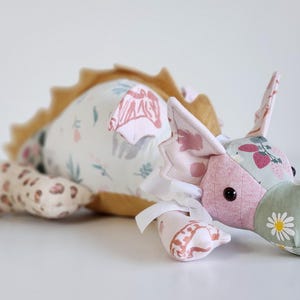 May include: A plush dragon toy crafted from various patterned fabrics, including a white body with floral prints, a sage green snout with a daisy pattern, and pink sections. It features a mustard yellow spine, small wings, and a white ribbon bow.