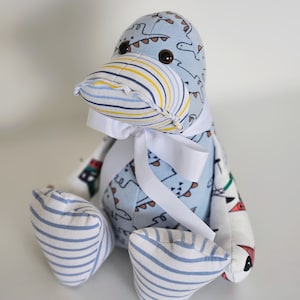 May include: A handmade penguin soft toy. The penguin is made from patterned fabric. The head and body are light blue with a dinosaur print. The beak has yellow, white, and blue stripes. The feet are blue and white striped. A white ribbon is tied around the neck.