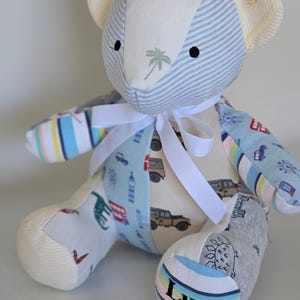 May include: A patchwork teddy bear with a white ribbon. The bear is made from various patterned fabrics, including blue stripes, palm tree prints, and vehicle designs. The bear's foot has the name "Luca" printed on it.