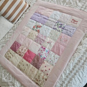 May include: A patchwork baby quilt in pink, lilac, and cream, with floral and patterned fabric squares. The quilt has a pink border and the name "Piper" and date "26.08.24" embroidered on it. A striped cushion is in the background.