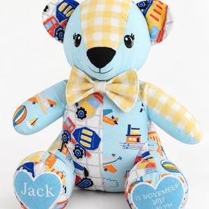 May include: A stuffed teddy bear made of light blue fabric with a yellow and white gingham pattern on the head, ears, and one arm. The body features a colourful print of construction vehicles and aeroplanes. A yellow gingham bowtie is at the neck.