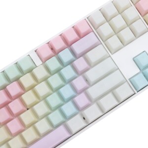 K&ouml;nnte beinhalten: Nahaufnahme einer wei&szlig;en mechanischen Tastatur mit pastellfarbenen Tastenkappen. Die Tastenkappen sind in einem Regenbogenverlauf angeordnet, der von Rosa &uuml;ber Gelb, Gr&uuml;n bis Blau reicht. Die Tastatur hat ein sauberes, modernes Design.