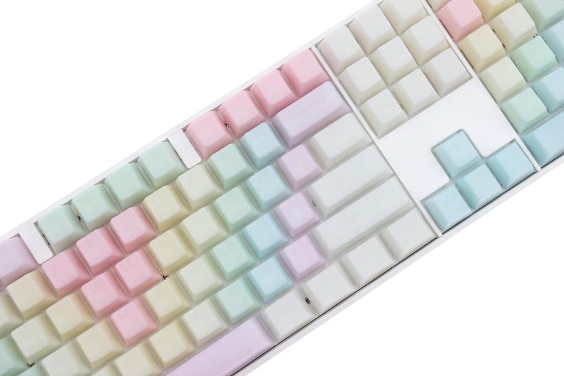 K&ouml;nnte beinhalten: Nahaufnahme einer wei&szlig;en mechanischen Tastatur mit pastellfarbenen Tastenkappen. Die Tastenkappen sind in einem Regenbogenverlauf angeordnet, der von Rosa &uuml;ber Gelb, Gr&uuml;n bis Blau reicht. Die Tastatur hat ein sauberes, modernes Design.