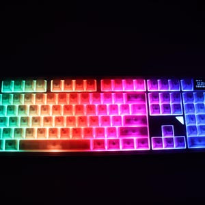 May include: A backlit computer keyboard with rainbow-colored keys. The keys transition from teal to red to purple, with a small display in the upper right corner. The keyboard is set against a black background.