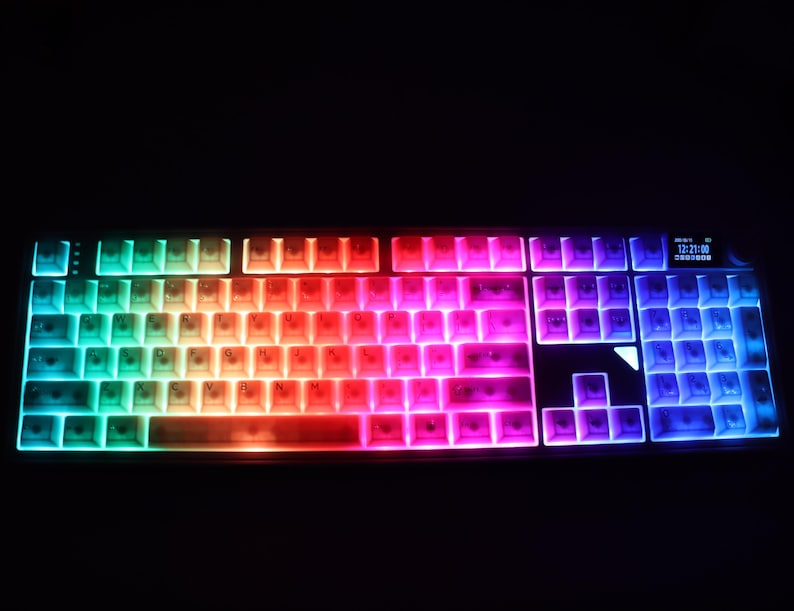 May include: A backlit computer keyboard with rainbow-colored keys. The keys transition from teal to red to purple, with a small display in the upper right corner. The keyboard is set against a black background.