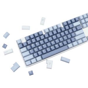 May include: A mechanical keyboard with a white frame and blue keycaps. The keycaps are a gradient of light and dark blue. Several keycaps are detached and scattered around the keyboard. The keyboard has a standard QWERTY layout.