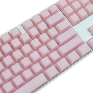 K&ouml;nnte beinhalten: Nahaufnahme einer mechanischen Tastatur mit hellrosa Tastenkappen. Die Tastatur hat einen silbernen Rahmen und ein Standard-QWERTZ-Layout. Die Tastenkappen sind einfarbig pastellrosa, und das Gesamtdesign ist sauber und modern.