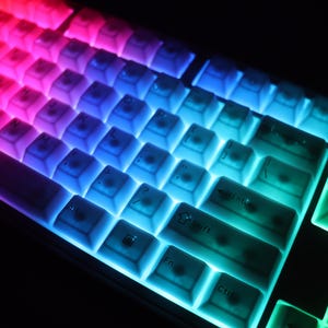 May include: Close-up of a backlit computer keyboard with a gradient of colors. The keys transition from red to purple, blue, and green. The keyboard is illuminated, creating a vibrant and colorful display.