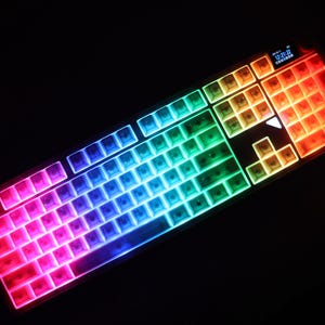 May include: A backlit computer keyboard with keys illuminated in a rainbow of colors. The keyboard has a black frame and a small display screen in the upper right corner. The keys are arranged in a standard QWERTY layout.