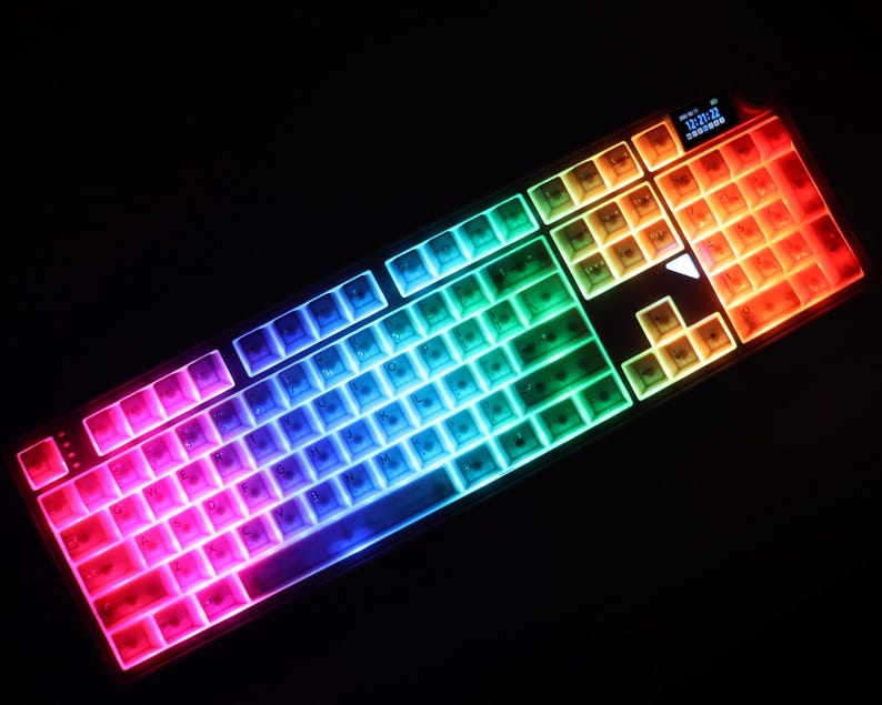 May include: A backlit computer keyboard with keys illuminated in a rainbow of colors. The keyboard has a black frame and a small display screen in the upper right corner. The keys are arranged in a standard QWERTY layout.
