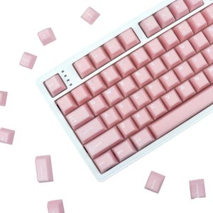 May include: A light pink mechanical keyboard with matching keycaps. The keyboard has a white frame and a standard QWERTY layout. Several keycaps are scattered around the keyboard. The keys are labeled with white lettering.