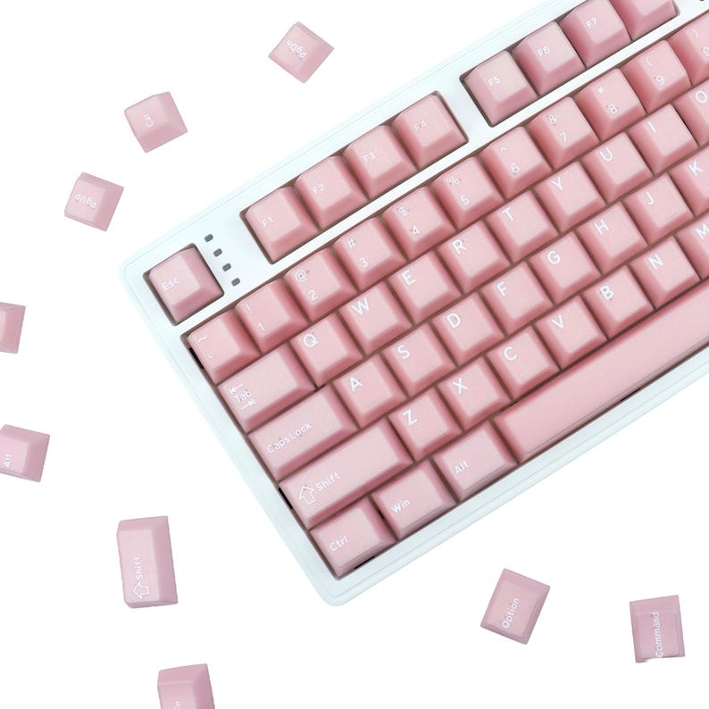 May include: A light pink mechanical keyboard with matching keycaps. The keyboard has a white frame and a standard QWERTY layout. Several keycaps are scattered around the keyboard. The keys are labeled with white lettering.