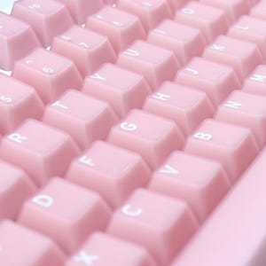May include: Close-up of a pink mechanical keyboard with white lettering on the keys. The keys are a soft, pastel pink color. The keyboard is angled, showing the keys in rows.