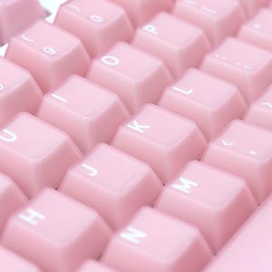 May include: Close-up of a computer keyboard with light pink keycaps. The keys are arranged in rows and columns, with white lettering and symbols. The keyboard has a clean, modern aesthetic.