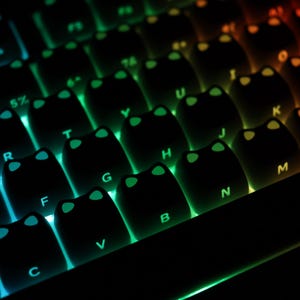 May include: Close-up of a backlit mechanical keyboard with black keycaps. The keys are illuminated with a gradient of colors, transitioning from blue to green and orange. The keycaps have a unique cat-ear design.