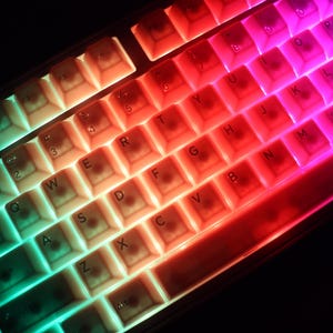 May include: A close-up of a mechanical keyboard with illuminated keys. The keys are lit with a gradient of colors, transitioning from green to red to purple. The keyboard has a standard QWERTY layout.