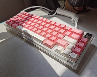 May include: A pink and white mechanical keyboard with a clear plastic case. The keyboard has a row of function keys at the top and a row of arrow keys at the bottom. The keys are labeled with letters, numbers, and symbols.