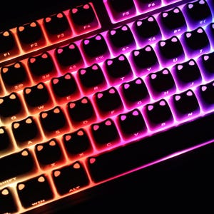 May include: Close-up of a mechanical keyboard with black keycaps featuring a cat face design. The keys are illuminated with a gradient of colors, transitioning from orange to pink and blue. The keyboard is designed for gaming or typing.