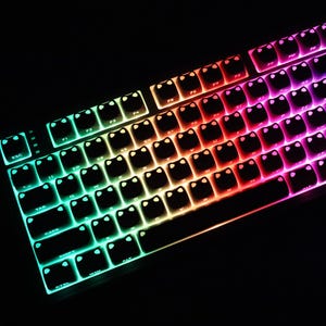 May include: A backlit mechanical keyboard with a black frame. The keys are illuminated with a rainbow gradient of colors, transitioning from green to red to purple. The keys have a unique cat face design.