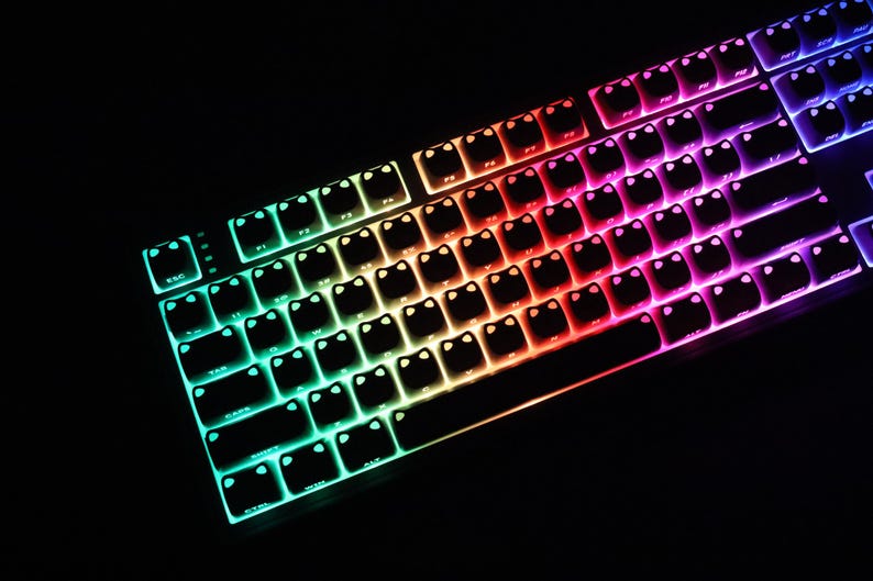 May include: A backlit mechanical keyboard with a black frame. The keys are illuminated with a rainbow gradient of colors, transitioning from green to red to purple. The keys have a unique cat face design.