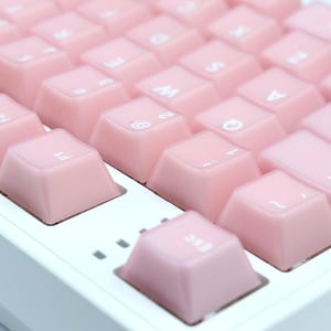May include: Close-up of a mechanical keyboard with light pink keycaps. The keys are arranged in a grid pattern, with white lettering visible on some keys. The keyboard has a white base and a minimalist design.