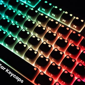 May include: Close-up of a keyboard with black keycaps shaped like cat ears. The keys are backlit with a gradient of colors, transitioning from blue to green, yellow, orange, and red. The text "Cute Cat Ear Keycaps" is visible.