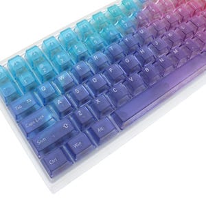 May include: A gradient-colored mechanical keyboard with transparent keycaps. The colors transition from blue to purple to pink. The keyboard has a standard layout with letters, numbers, and function keys. The keycaps are clear, allowing the internal components to be visible.
