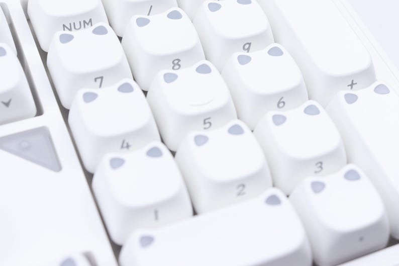 May include: Close-up of a white keyboard with custom keycaps. The keycaps are white with a light gray frog-like design. The number keys are visible, and the "NUM" key is also visible.