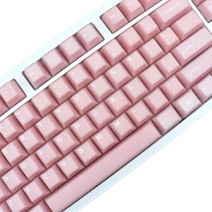May include: A close-up of a mechanical keyboard with pink keycaps and a white frame. The keys are arranged in a standard QWERTY layout, with function keys and number keys visible. The keyboard has a clean, modern aesthetic.