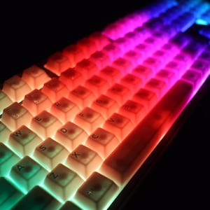 May include: A close-up of a mechanical keyboard with illuminated keys. The keys are lit with a gradient of colors, transitioning from green to red to blue. The keyboard has a compact layout with white keycaps.