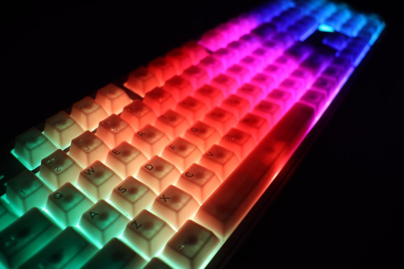 May include: A close-up of a mechanical keyboard with illuminated keys. The keys are lit with a gradient of colors, transitioning from green to red to blue. The keyboard has a compact layout with white keycaps.