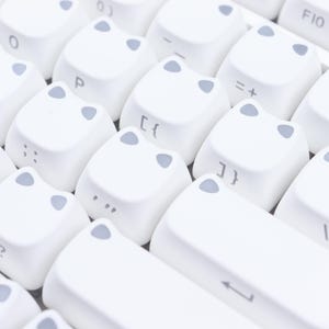 May include: Close-up of a white keyboard with cat-shaped keycaps. The keycaps have gray accents and feature letters, numbers, and symbols. The keyboard is designed for a unique and playful aesthetic.