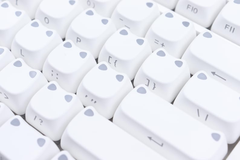 May include: Close-up of a white keyboard with cat-shaped keycaps. The keycaps have gray accents and feature letters, numbers, and symbols. The keyboard is designed for a unique and playful aesthetic.