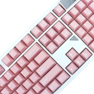 May include: A close-up of a white and pink mechanical keyboard. The keyboard has pink keycaps with white lettering and a white frame. The keys include function keys, number keys, and arrow keys.