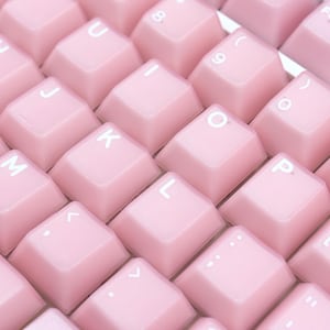 May include: Close-up of a pink mechanical keyboard. The keycaps are a soft pastel pink with white lettering. The keys are arranged in a grid pattern, with the letters and numbers clearly visible. The keyboard has a clean, modern aesthetic.