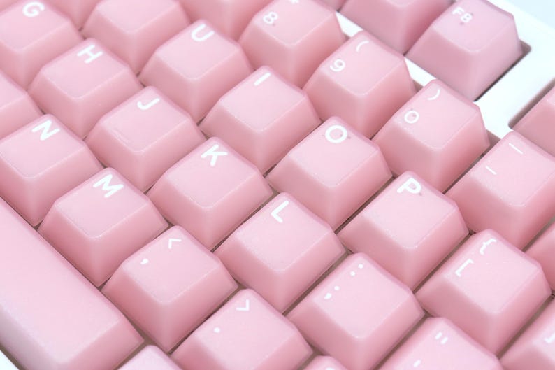 May include: Close-up of a pink mechanical keyboard. The keycaps are a soft pastel pink with white lettering. The keys are arranged in a grid pattern, with the letters and numbers clearly visible. The keyboard has a clean, modern aesthetic.