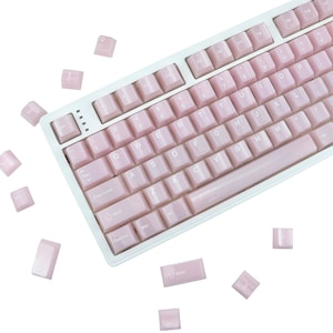 May include: A white mechanical keyboard with pink keycaps. The keyboard has a standard layout with alphanumeric keys, function keys, and a space bar. Several pink keycaps are scattered around the keyboard. The keys are labeled with white lettering.