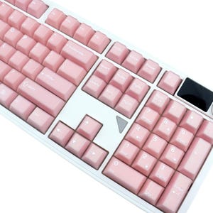 May include: A full-sized computer keyboard with a white frame and light pink keycaps. The keys are labeled with white lettering. The keyboard has a number pad and function keys. The design is clean and modern.