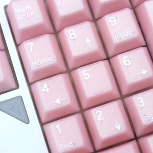May include: Close-up of a computer keyboard with light pink keys. The keys are arranged in a grid pattern and feature white lettering and symbols. The number pad keys are visible, with numbers and directional arrows.