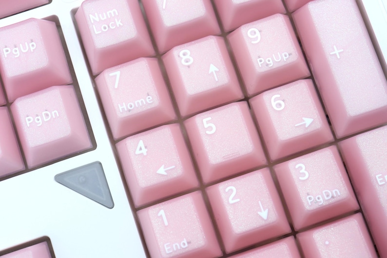 May include: Close-up of a computer keyboard with light pink keys. The keys are arranged in a grid pattern and feature white lettering and symbols. The number pad keys are visible, with numbers and directional arrows.