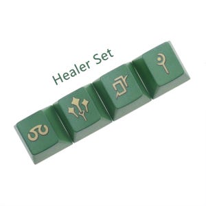 Puede incluir: Un conjunto de cuatro teclas verdes con s&iacute;mbolos dorados. Las teclas est&aacute;n etiquetadas como "Healer Set" y presentan s&iacute;mbolos que representan la magia de curaci&oacute;n.