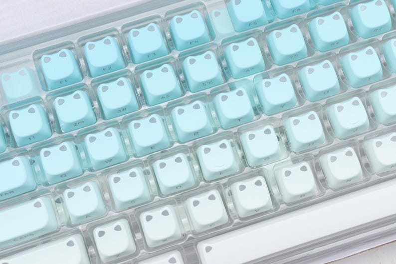 May include: A close-up of a computer keyboard with light blue and white keycaps. Each keycap has a frog face design. The keycaps are arranged in rows and columns, with a gradient effect from light blue to white.