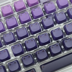 May include: A close-up of a keyboard with custom keycaps. The keycaps are a gradient of purple hues, each featuring a stylized frog face. The keyboard keys are arranged in a grid pattern, with a dark purple frame.